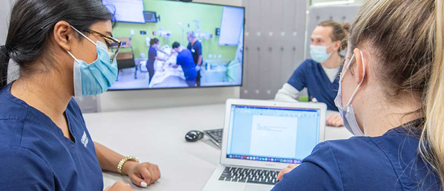 Two healthcare professionals with masks discussing work in front of a laptop, with a screen in the background displaying an ongoing medical training or simulation session.